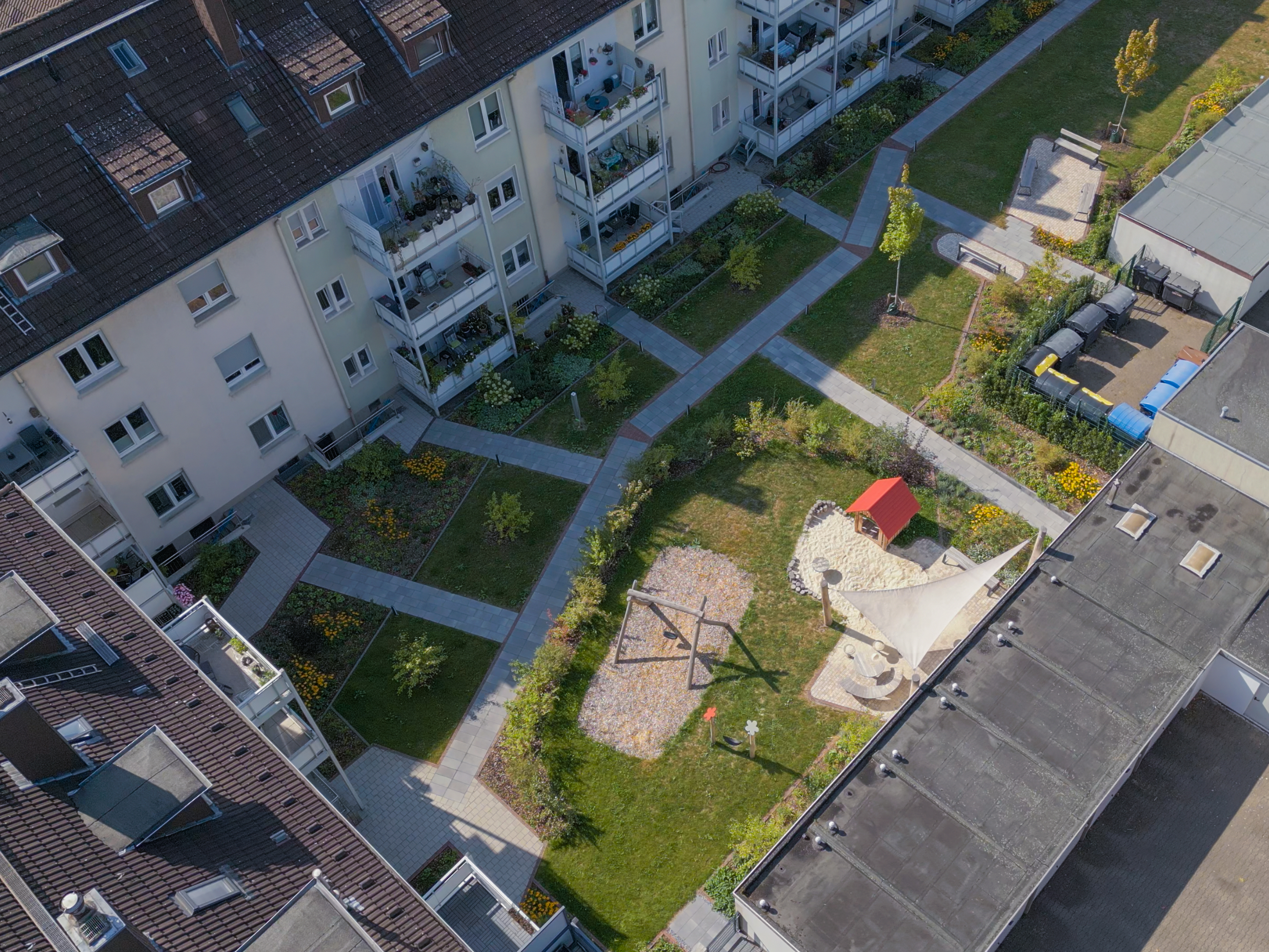 Luftaufnahme eines begrünten Innenhofs mit Wegen, Rasenflächen, Spielbereich und umliegenden Mehrfamilienhäusern mit Balkonen.