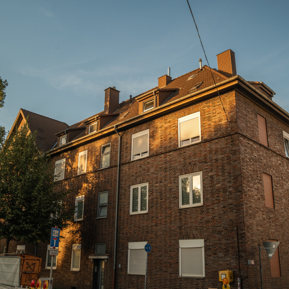 Ein mehrgeschossiges Backsteingebäude mit weißen Fenstern steht an einer Straßenecke und wird vom warmen Abendlicht beleuchtet.