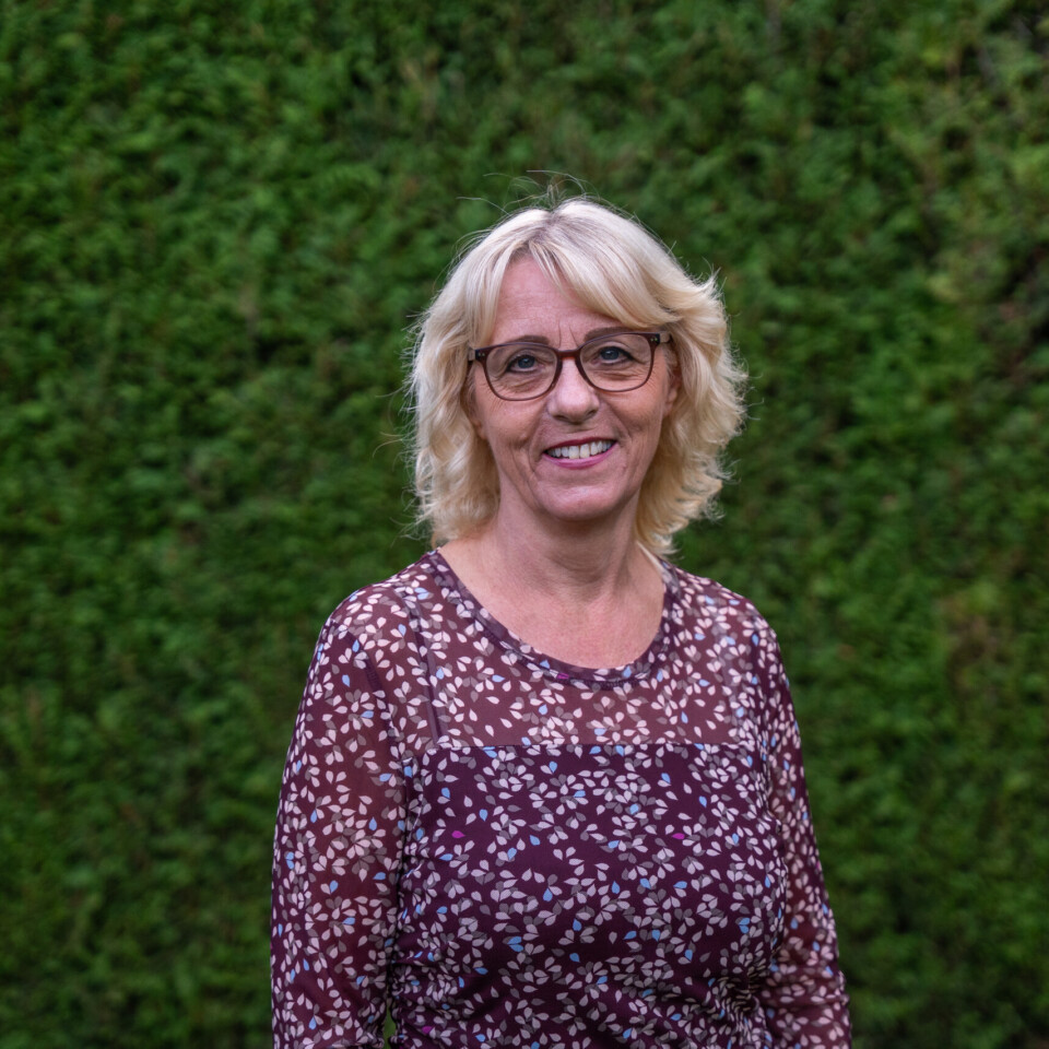 Eine Frau mit blonden Haaren, Brille und gemusterter Bluse steht lächelnd vor einer grünen Hecke.