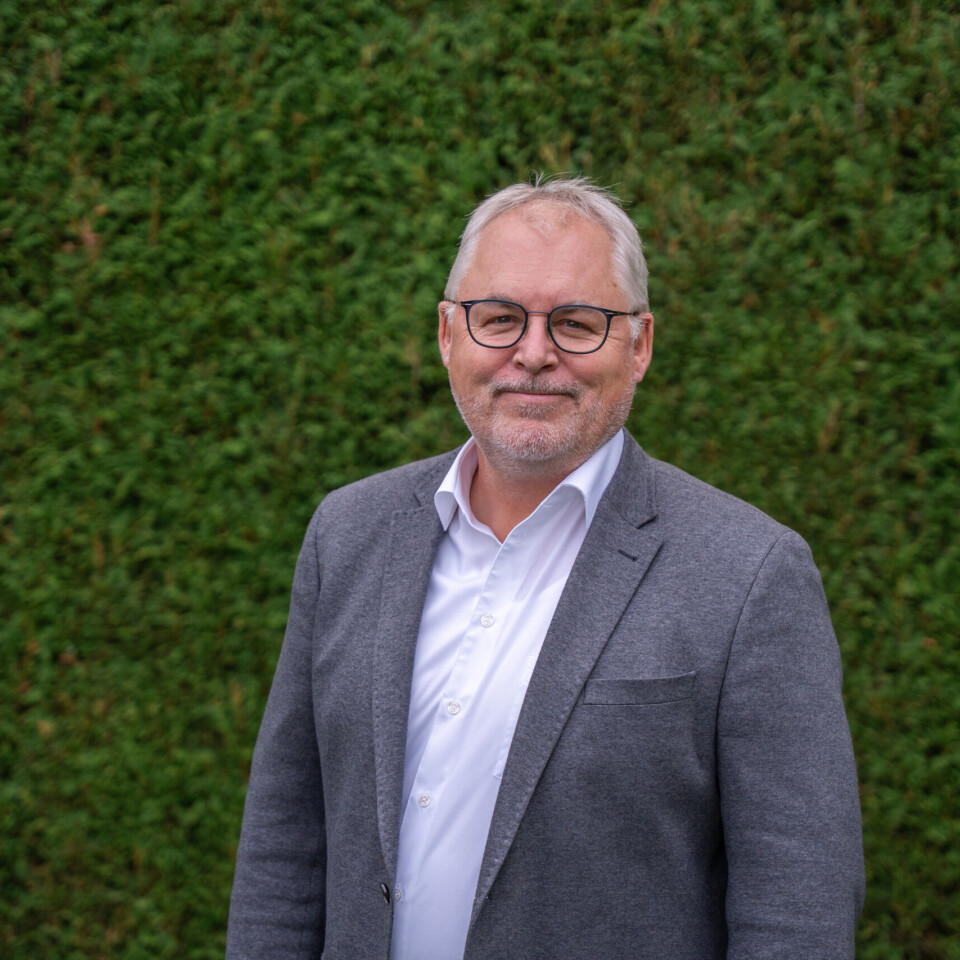 Ein Mann mittleren Alters mit Brille, weißem Hemd und grauem Sakko steht lächelnd vor einer dichten grünen Hecke.