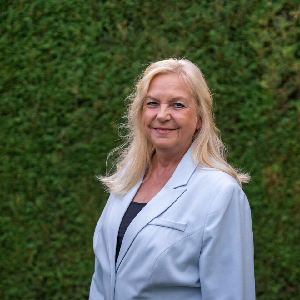 Eine Frau mit langen blonden Haaren trägt ein helles Sakko und steht lächelnd vor einer dichten grünen Hecke.