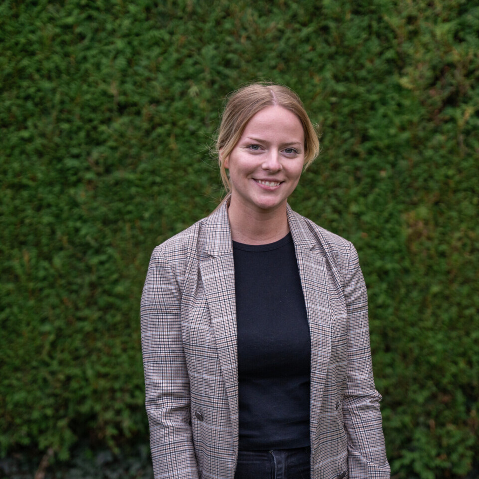 Lächelnde Mitarbeiterin der Heimbau Herne im karierten Blazer vor grüner Hecke – Halbporträt im Freien.