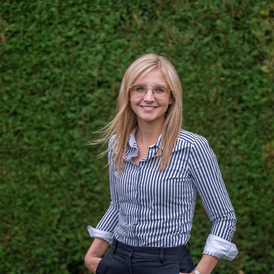 Eine Frau mit blonden Haaren, Brille und gestreiftem Hemd steht lächelnd vor einer grünen Hecke.