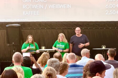 Drei Personen stehen auf der Bühne. Im Vordergrund sind mehrere Menschen im Publikum zu sehen