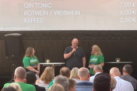 Drei Personen stehen auf der Bühne. Im Vordergrund sind mehrere Menschen im Publikum zu sehen