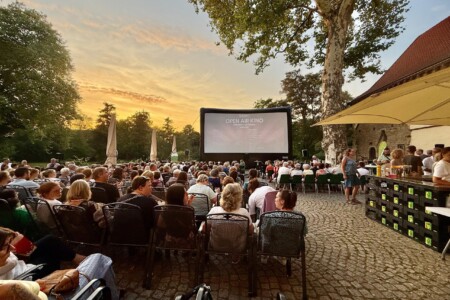 Viele Menschen sitzen beim Sonnenuntergang im Freien und schauen auf eine große Kinoleinwand
