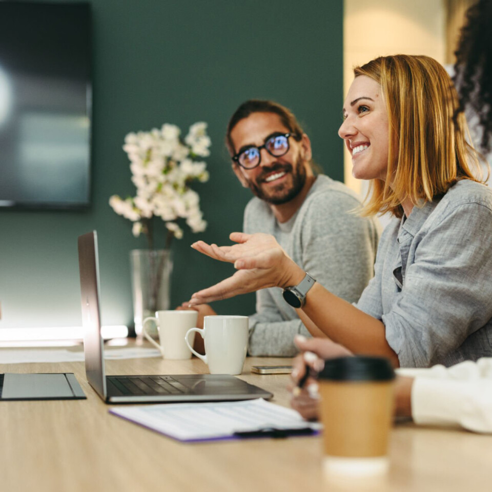 Mehrere Personen sitzen gemeinsam an einem Konferenztisch, sprechen miteinander und arbeiten mit Laptops und Notizen in einer hellen, modernen Büroumgebung.