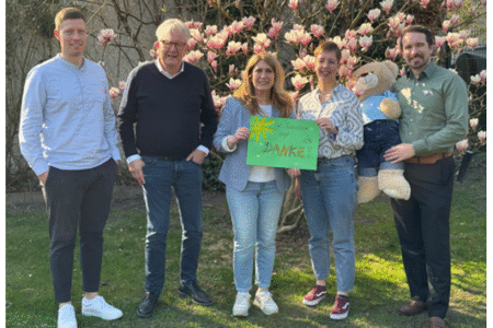Mehrere Personen stehen in einem Garten und posieren fürs Foto. Zwei Perosnen halten zusammen ein bemaltes Plakat hoch. Eine andere Person trägt einen großen Teddybär