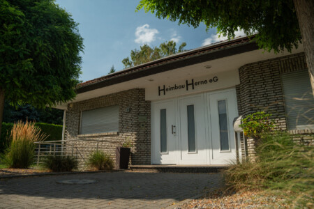 Außenaufnahme eines einstöckigen Hauses mit weißer Eingangstür, gemauerten Fassadesteinen und gepflegter Bepflanzung im Vordergrund.