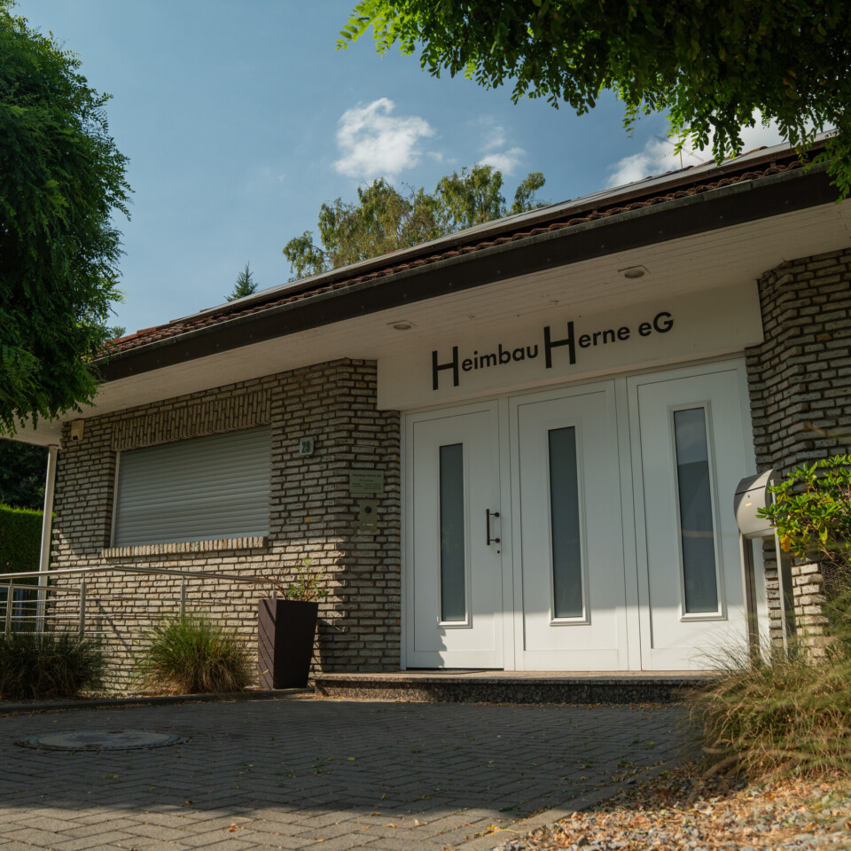 Außenaufnahme eines einstöckigen Hauses mit weißer Eingangstür, gemauerten Fassadesteinen und gepflegter Bepflanzung im Vordergrund.