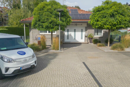Außenaufnahme des Gebäudes der Heimbau Herne eG mit weißem Eingangsbereich, zwei Bäumen und einem Firmenfahrzeug auf dem Parkplatz.