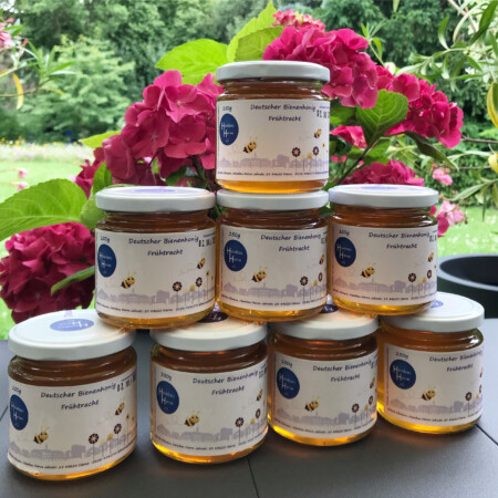 Mehrere Gläser mit Honig der Heimbau Herne eG sind pyramidenförmig gestapelt. Im Hintergrund blühen pinke Hortensien und grüne Gartenpflanzen.
