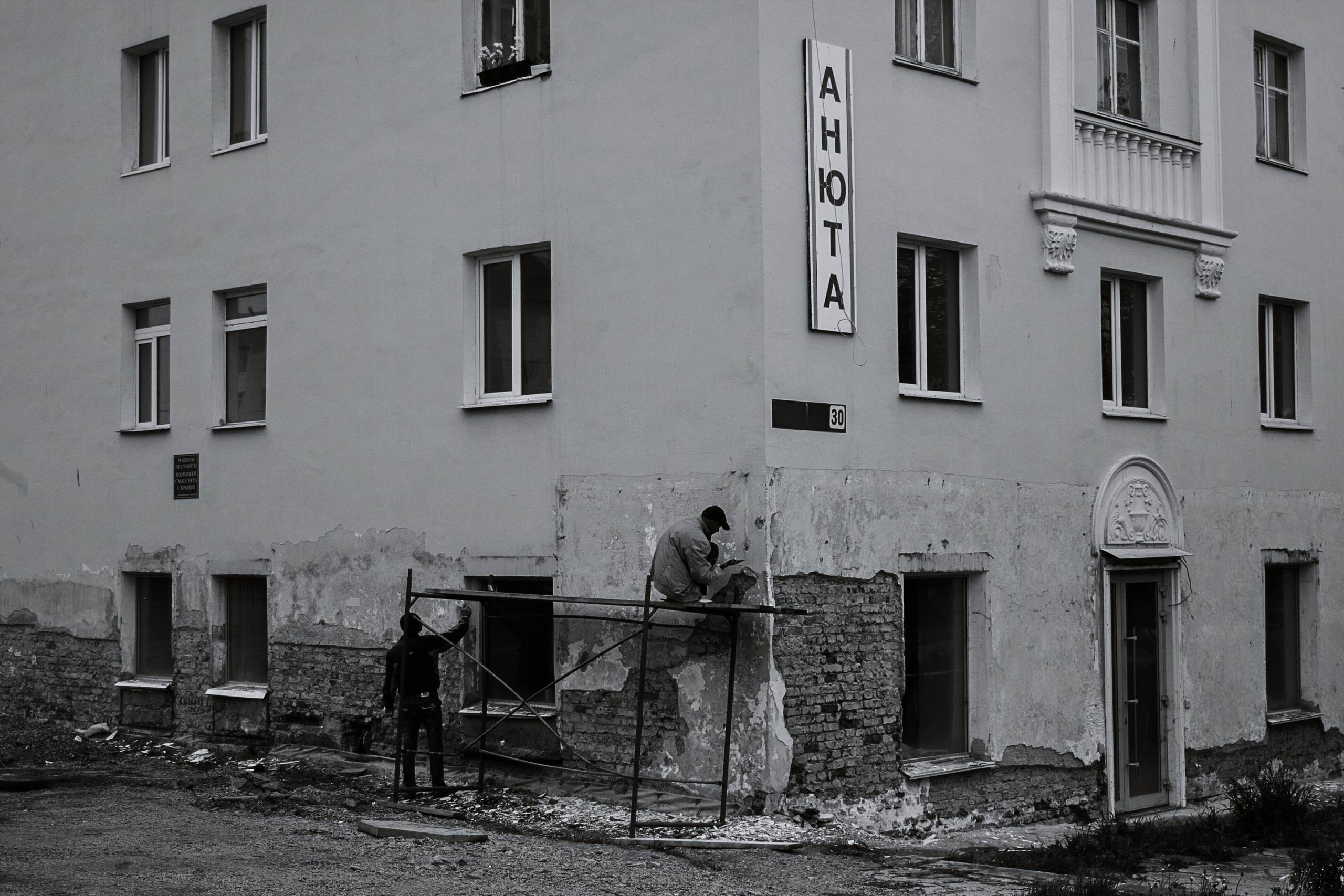 Schwarz-weiß-Aufnahme eines Wohngebäudes, an dessen Fassade zwei Arbeiter auf einem Gerüst Ausbesserungsarbeiten durchführen.
