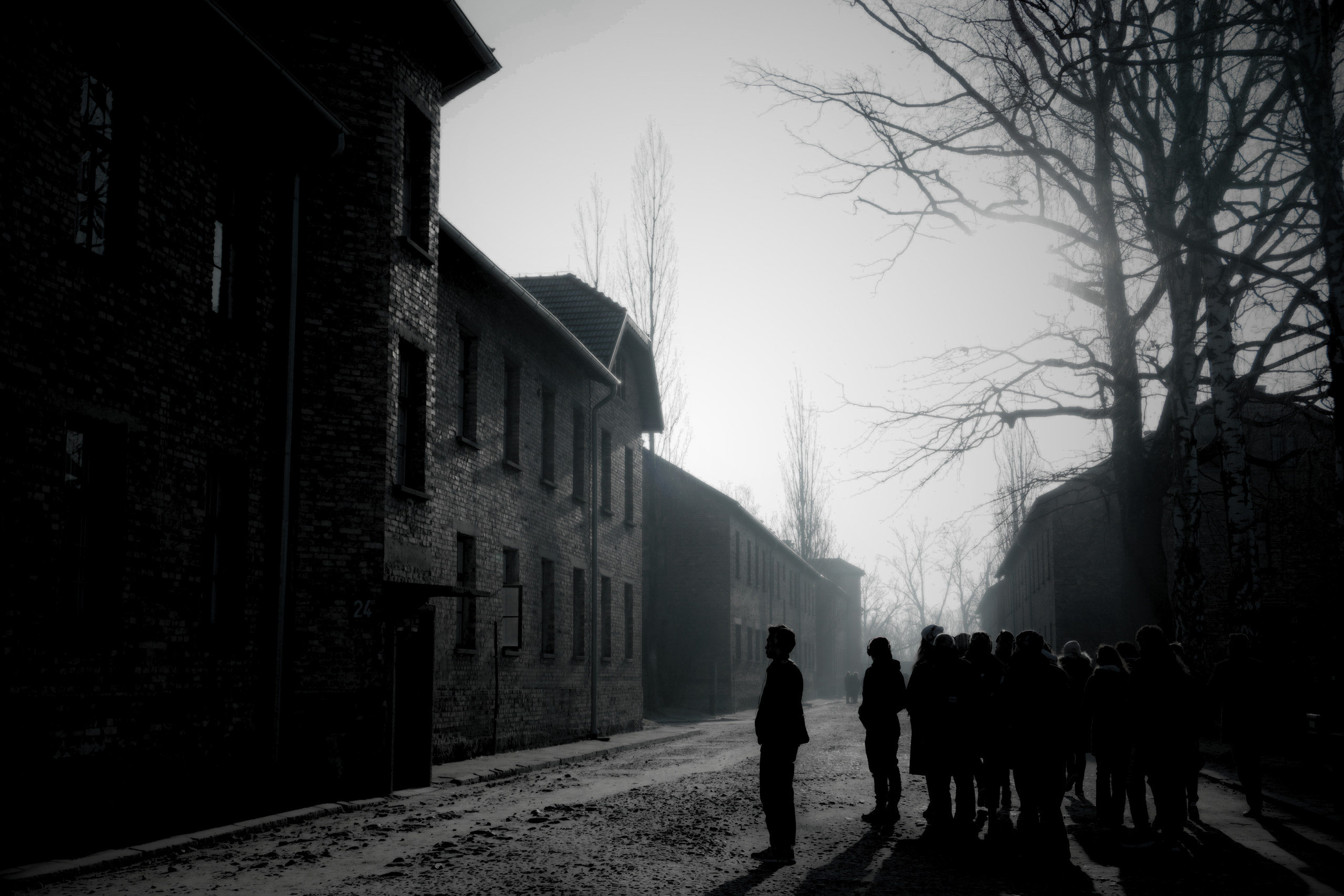 Schwarz-weiß-Fotografie eines historischen Gebäudekomplexes aus Backstein. Eine Gruppe von Menschen steht im Vordergrund auf einer Straße, die von kahlen Bäumen gesäumt ist.