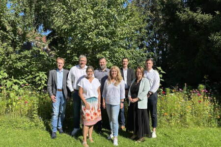 Acht Personen stehen in einer sonnigen Gartenlandschaft vor Bäumen und Blumen und posieren für ein Gruppenfoto.