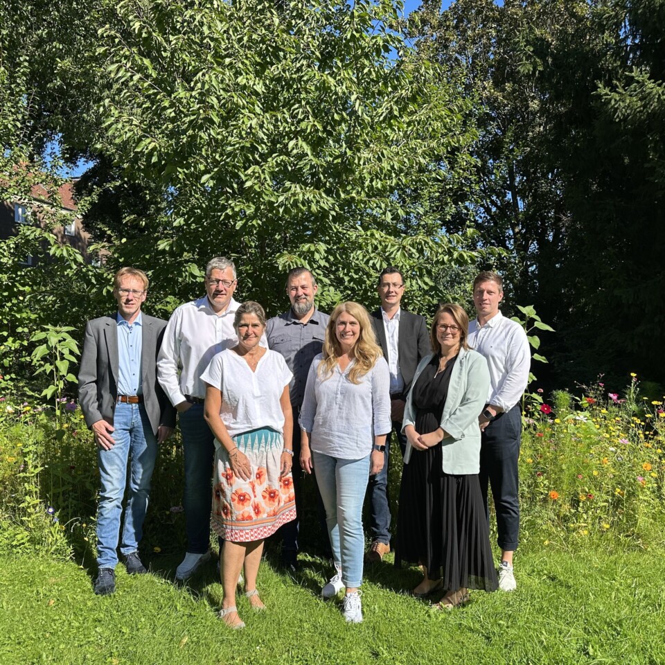 Acht Personen stehen in einer sonnigen Gartenlandschaft vor Bäumen und Blumen und posieren für ein Gruppenfoto.