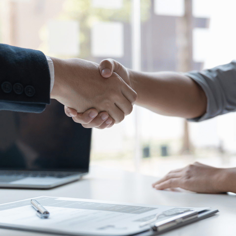 Zwei Personen geben sich bei einem Gespräch im Büro die Hand, auf dem Tisch liegen Dokumente und ein Laptop.
