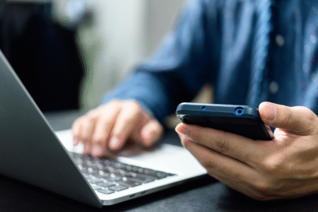 Eine Person bedient ein Smartphone in der Hand, während die andere Hand auf der Tastatur eines geöffneten Laptops liegt.