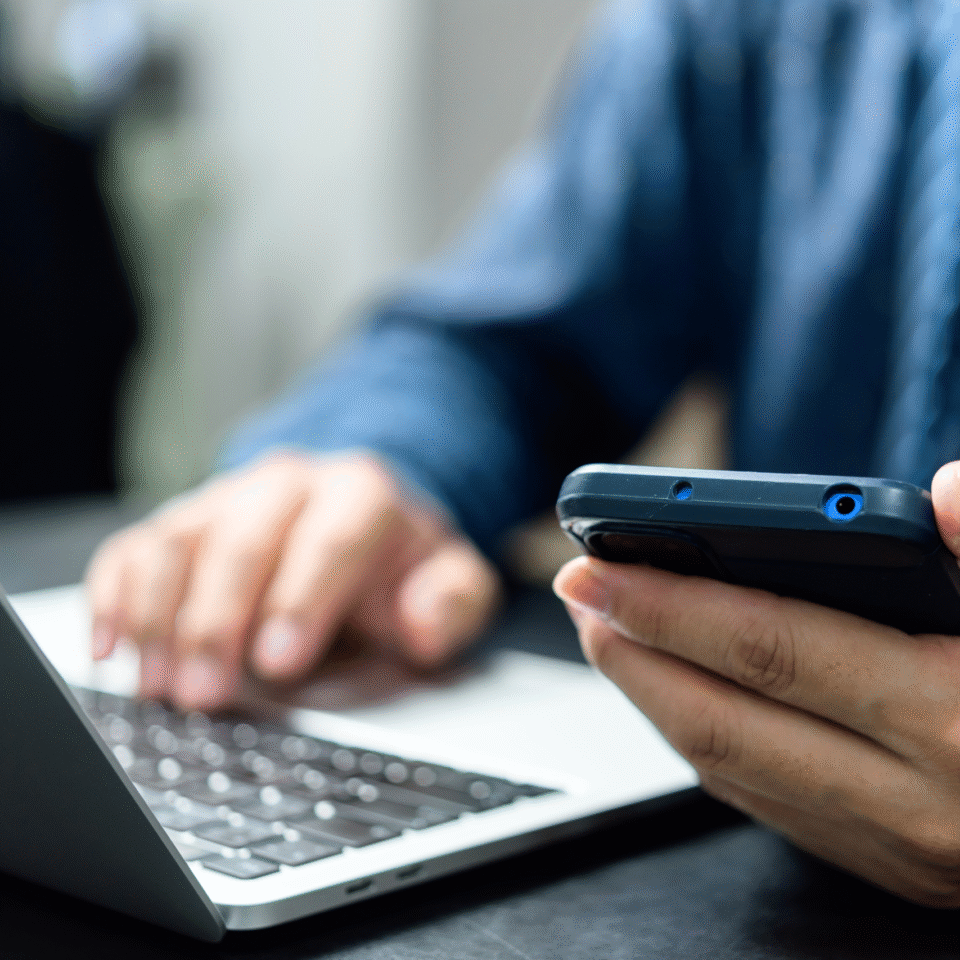 Eine Person bedient ein Smartphone in der Hand, während die andere Hand auf der Tastatur eines geöffneten Laptops liegt.