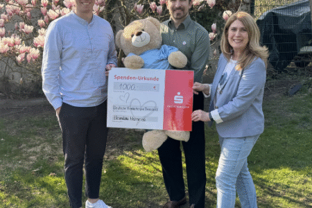 Drei Personen halten einen Spendencheck in der Hand. Die Person in der mitte dazu noch einen großen Teddybär