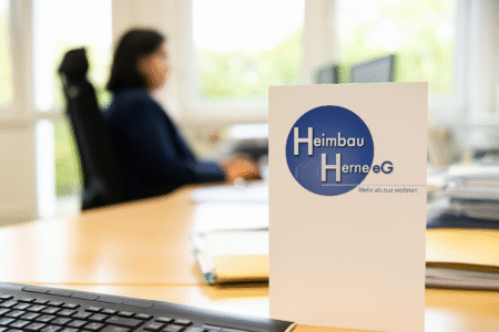 Broschüre der Heimbau Herne eG auf einem Schreibtisch, im Hintergrund sitzt eine Mitarbeiterin am Computer.