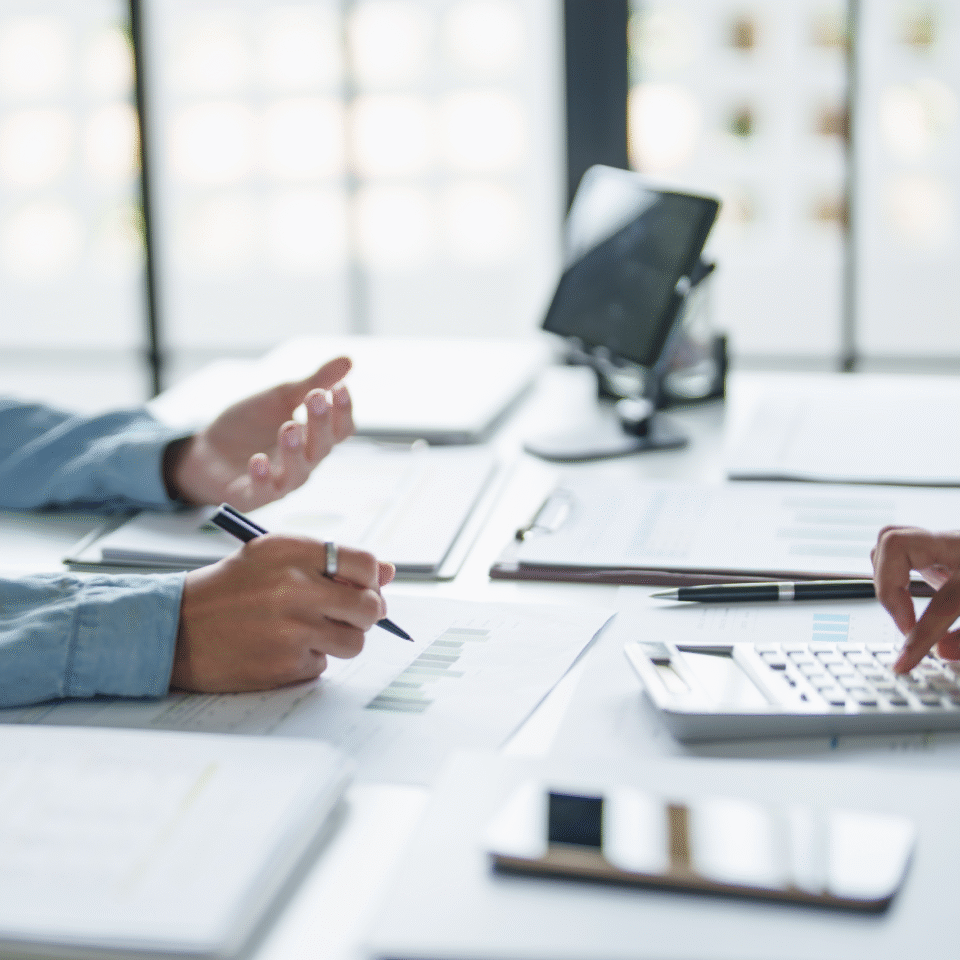 Zwei Personen sitzen in einem Büro am Schreibtisch. Die eine Person schreibt etwas auf, die andere arbeitet mit einem Taschenrechner