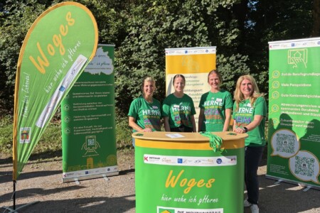 vier Frauen stehen auf einer Berufsmesse an ihrem Stand. Der Stand und die Klamotten der Personen sind grün