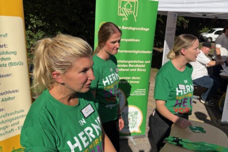Frauen stehen auf einer Berufsmesse an ihrem Stand. Der Stand und die Klamotten der Personen sind grün