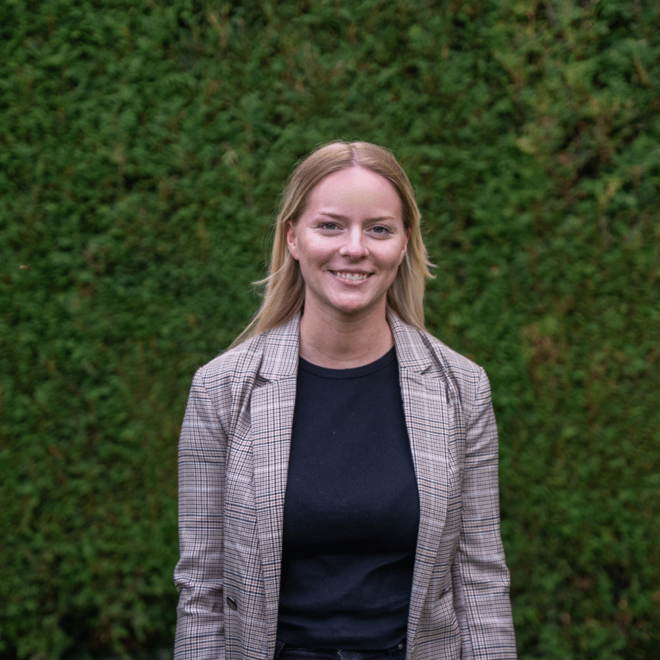 Lächelnde Mitarbeiterin der Heimbau Herne im karierten Blazer vor grüner Hecke – Halbporträt im Freien.