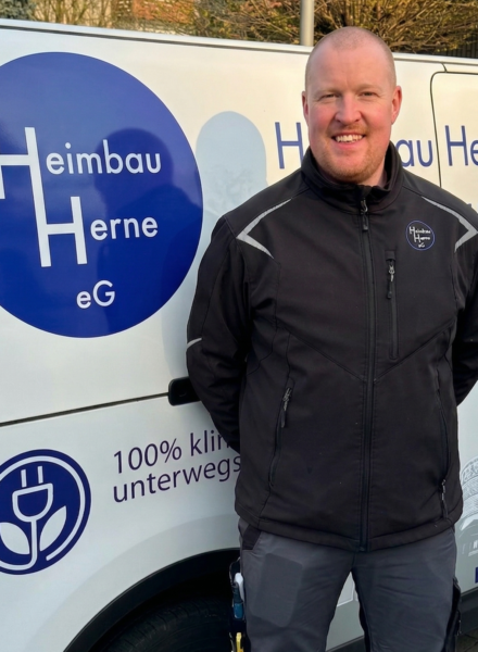 Ein Mann in dunkler Arbeitsjacke steht vor einem Firmenfahrzeug mit dem Logo der Heimbau Herne eG.
